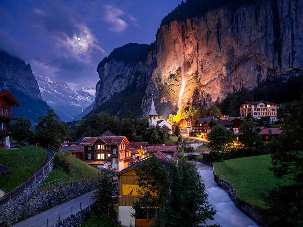 A twilight view of Lauterbrunnen Valley, featuring traditional wooden chalets, a cascading waterfall, and towering cliffs illuminated by soft moonlight.