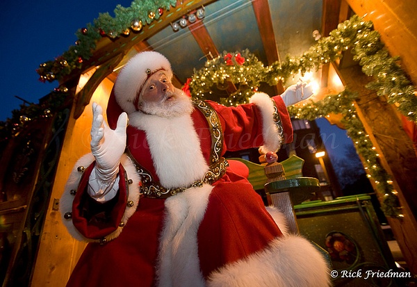 Santa Claus waving, hanging off a cart  with Christmas lights at dusk by Rick Friedman - Portraits - Rick Friedman Photography 