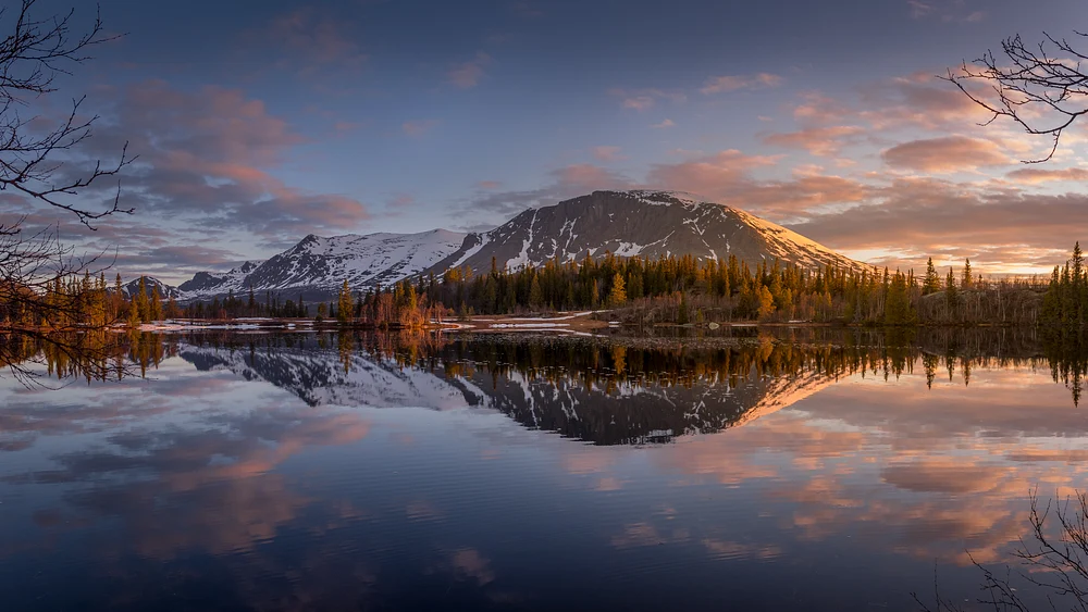 A tranquil spring scene at Skogshorn Mountain, featuring a serene lake reflecting the majestic peaks and colorful sunrise sky.