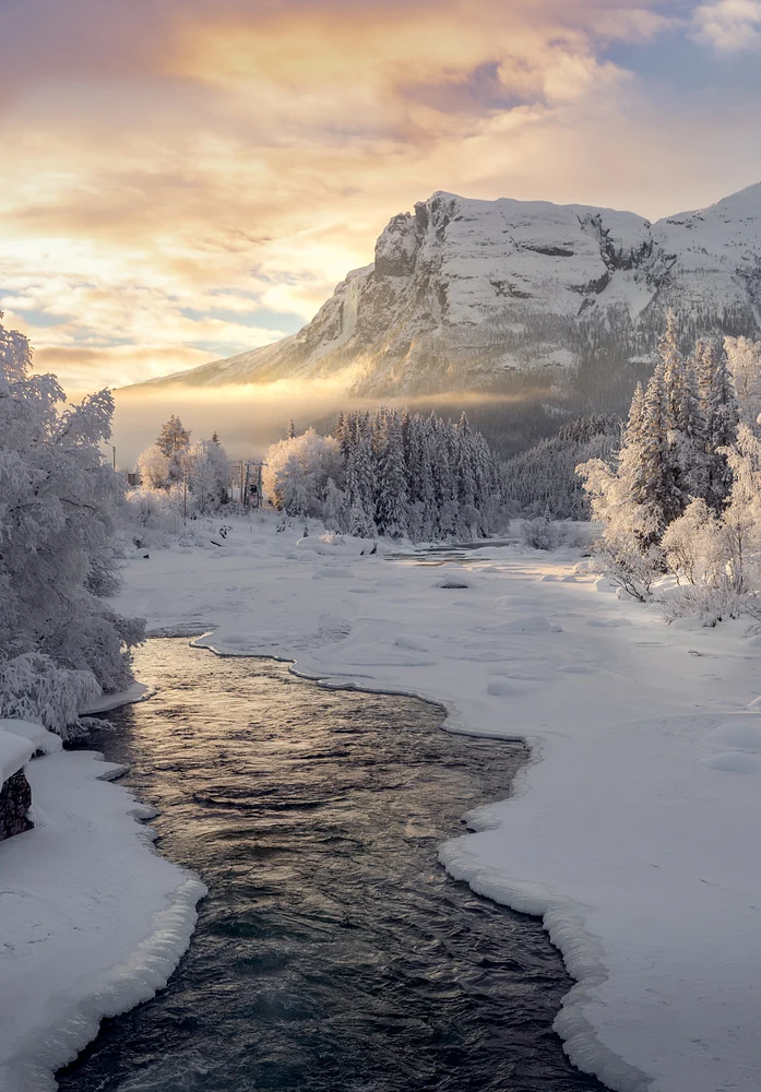 A serene winter landscape at Veslehodn featuring a winding river surrounded by snow-covered trees and mountains, illuminated by a warm sunset glow.