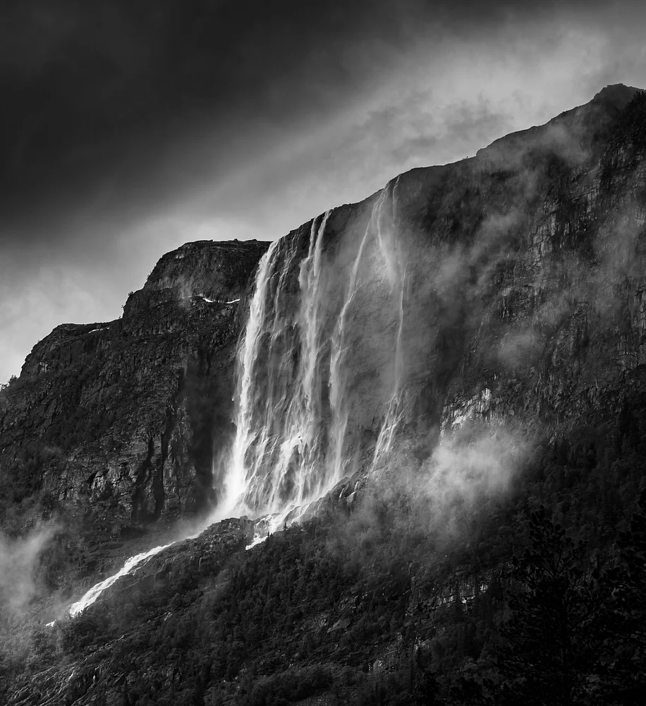 Black and white photograph of Hydnefossen Waterfall cascading down a rocky cliff, surrounded by mist and dramatic clouds.