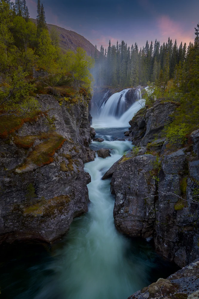 A stunning view of Rjukandefossen waterfall at midnight, surrounded by cliffs and lush greenery, with a tranquil river flowing beneath.