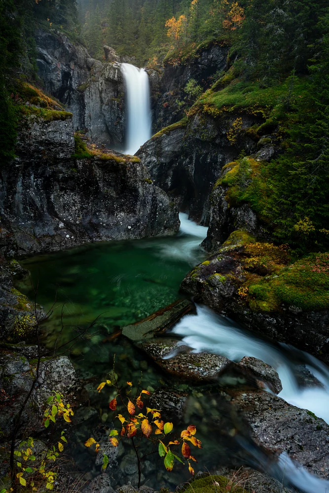 Tr&oslash;imsanifossen waterfall cascading into an emerald pool surrounded by autumn foliage and rocky terrain.