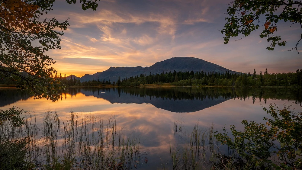 A stunning sunset over Skogshorn Mountain, reflecting on a calm lake surrounded by lush greenery and gentle grasses.