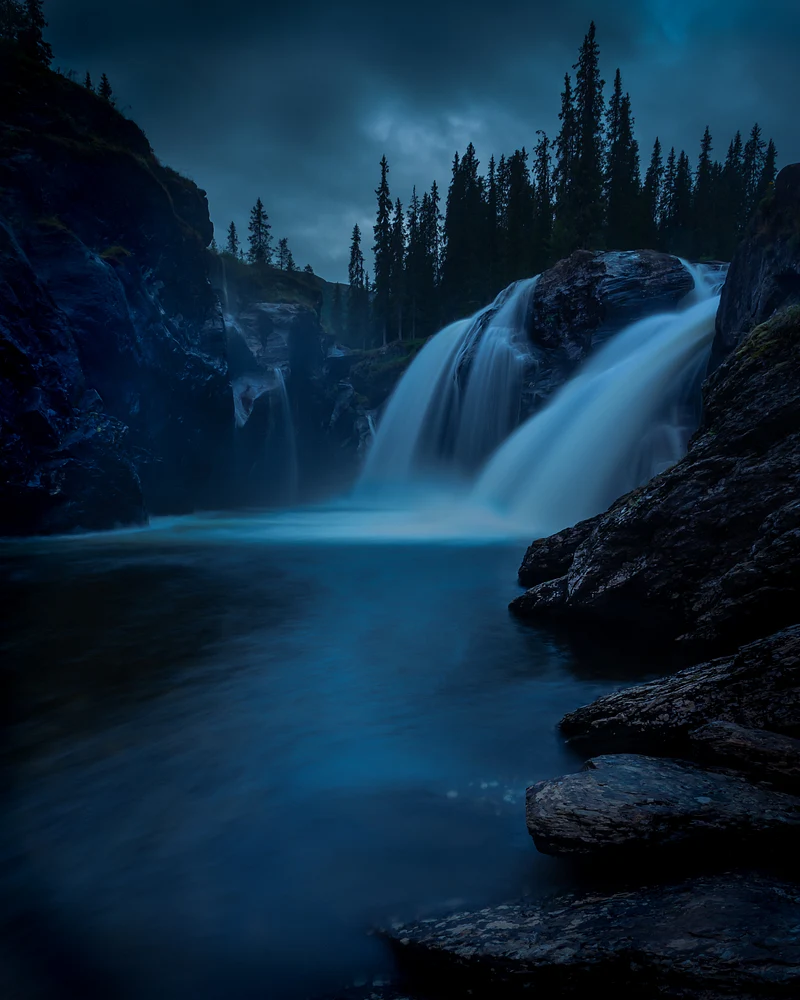 A captivating view of Rjukandefossen waterfall at dusk, surrounded by rocky cliffs and evergreen trees, showcasing the serene blue waters cascading down.