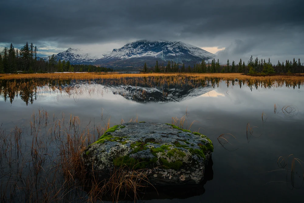 Reflection of Skogshorn mountain in a tranquil lake surrounded by autumn foliage and a moss-covered stone in the foreground.
