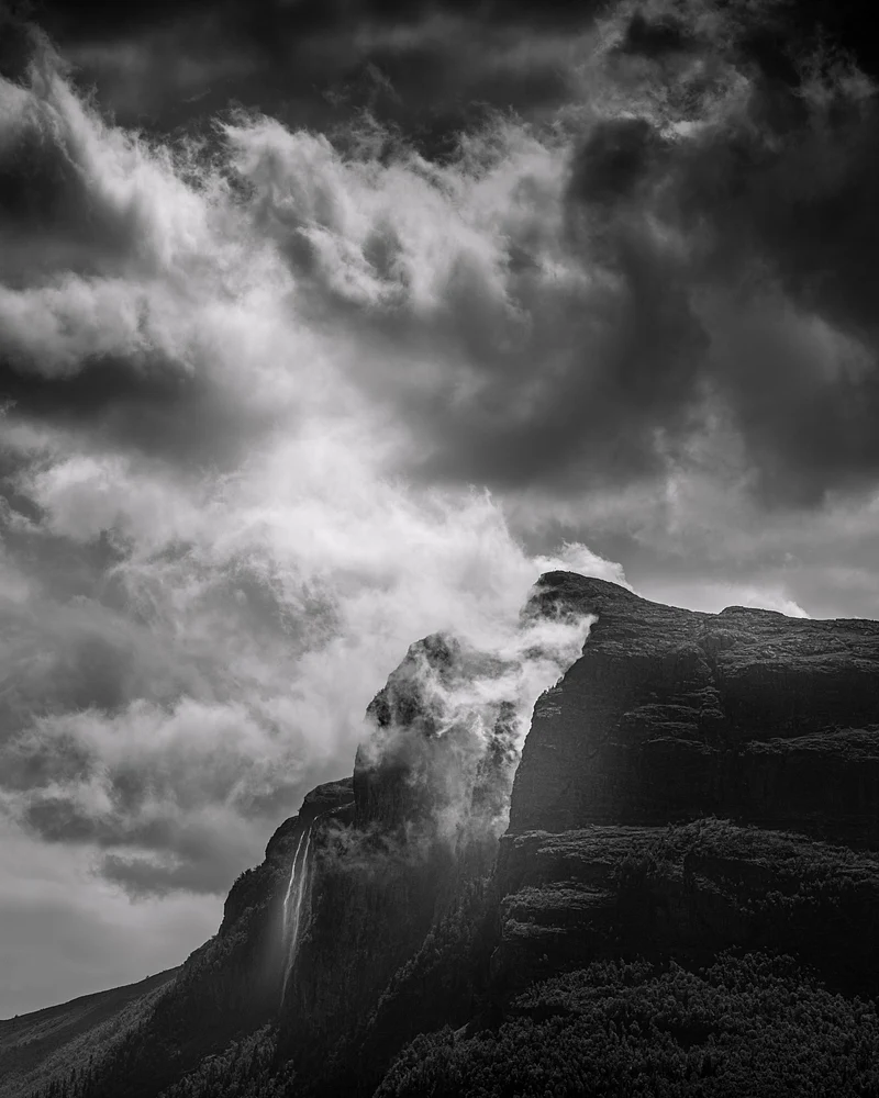 Black and white photograph of dramatic clouds over Vesleh&oslash;dn Mountain, showcasing mist and rugged peaks.