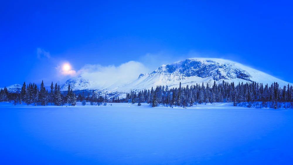 Full moon shining over Skogshorn Mountain with a snowy landscape and evergreen trees in the foreground.