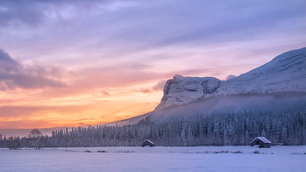 A serene winter sunset over snow-covered mountains and rustic cabins, with soft orange and purple hues in the sky.