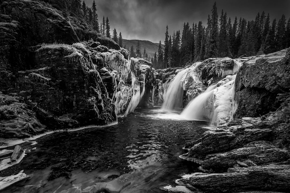 Black and white image of a waterfall cascading over rocks in a forest landscape, surrounded by trees and a moody sky.