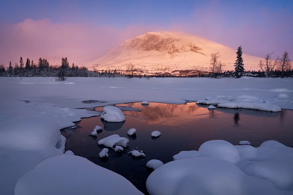 A serene winter landscape featuring a snow-covered mountain at sunrise, with soft pink and orange hues reflecting in a tranquil river surrounded by snow-laden rocks.