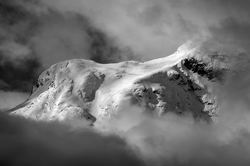 Black and white photograph of a majestic snow-capped mountain peak partially obscured by clouds.