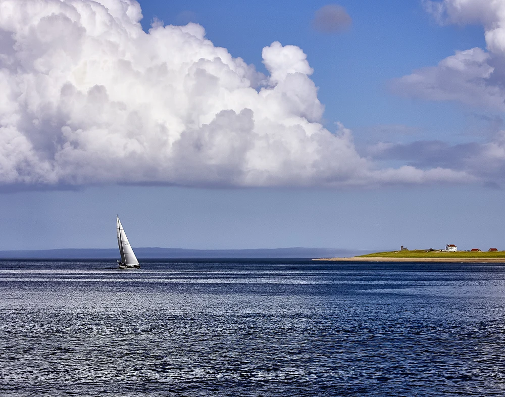 A sailboat gliding on calm Cape Cod waters under a blue sky with clouds.