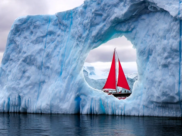 A vibrant red sailboat passes through an ice arch in an Arctic landscape.