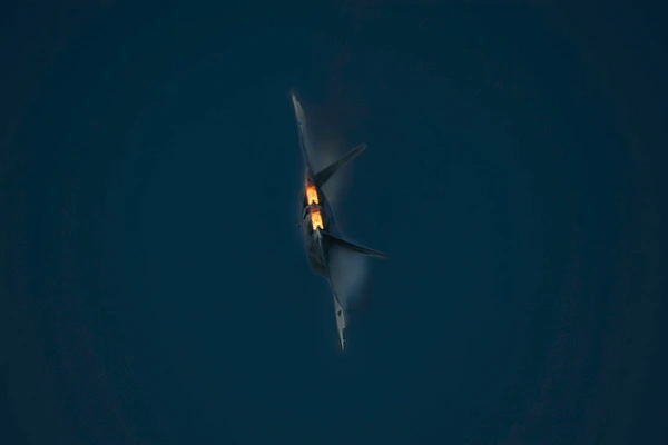 F-22 Raptor banking at an air show with bright twin afterburners and vapor trails against a dark blue sky